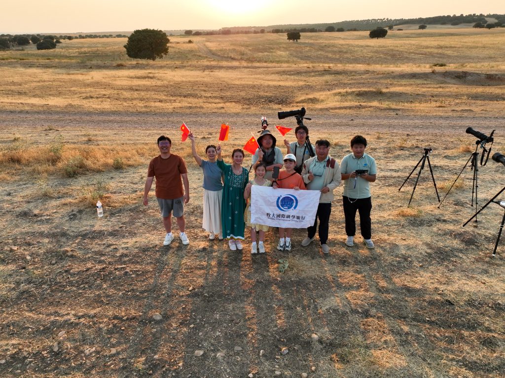 Nine members of the eclipse trip holding flags on golden grass at the Lerma site